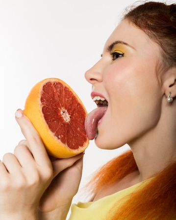 Young redhead woman licks a grapefruit on a white background. concept of healthy eatingの写真素材