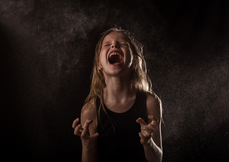 young girl with live emotions dancing under water drops. child screaming on a dark background with water splashの写真素材