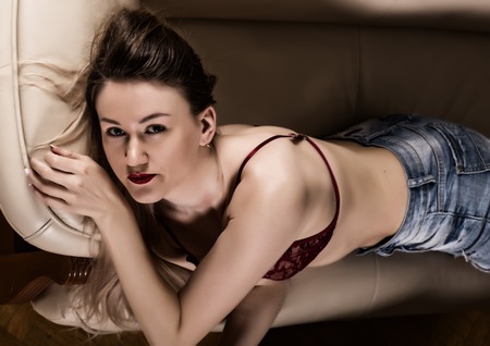 seductive young woman in red bra and jeans resting on sofa. sexy hipster girl enjoying on the couchの写真素材