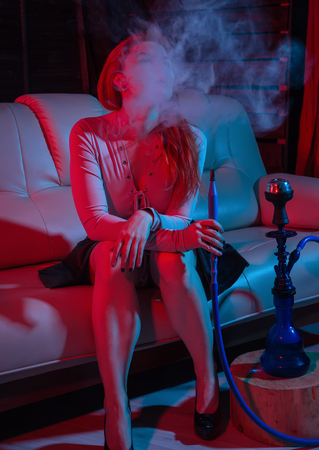 Sexy redhead woman vaping electronic hookah in red blue tonesの写真素材