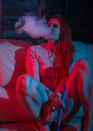 Sexy redhead woman vaping electronic hookah in red blue tonesの写真素材