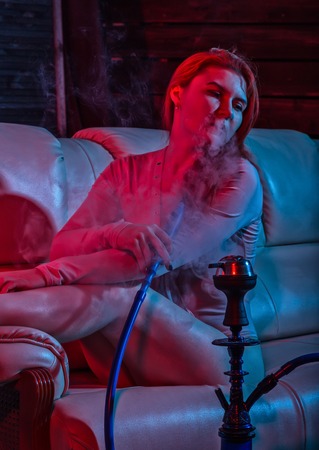 Sexy redhead woman vaping electronic hookah in red blue tones. girl sitting on a sofa and blows white doomの写真素材