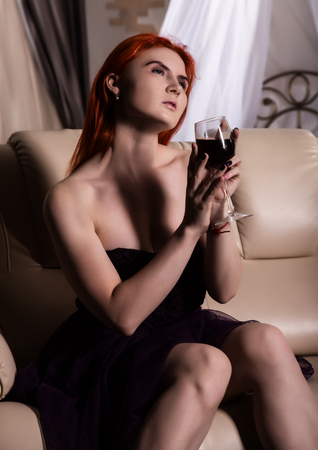 lonely young woman drinking wine in hotel room. redhead woman sitting on a sofa with glass of red wineの写真素材