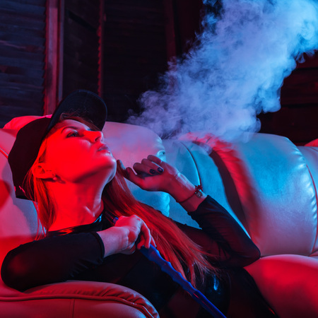 sexy redhead woman in black bodysuit sitting on a sofa and smokes hookah, girl blows out big smoke, red blue tonesの写真素材
