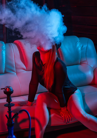 sexy redhead woman in black bodysuit sitting on a sofa and smokes hookah, girl blows out big smoke, red blue tonesの写真素材