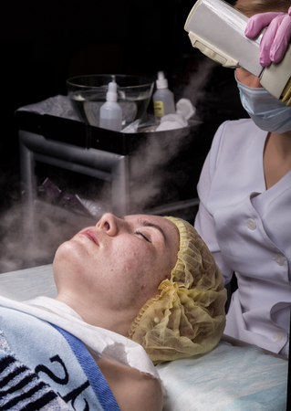 Doctor cleanses the womans skin with a steam. young woman with problem skin at the beautician. Cosmetology conceptの写真素材