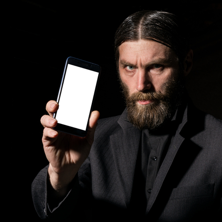 Old believer senior priest with a smartphone, bearded old man is calling on a dark backgroundの写真素材