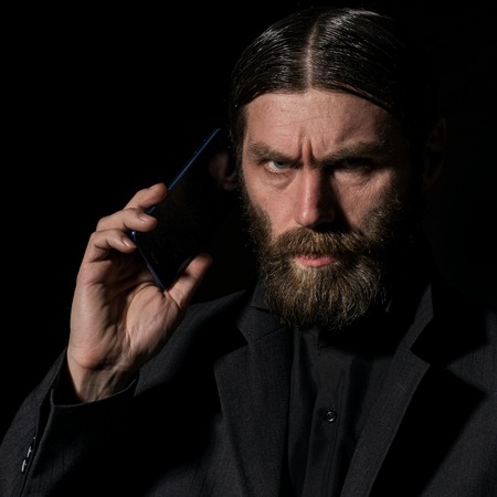 Old believer senior priest with a smartphone, bearded old man is calling on a dark backgroundの写真素材