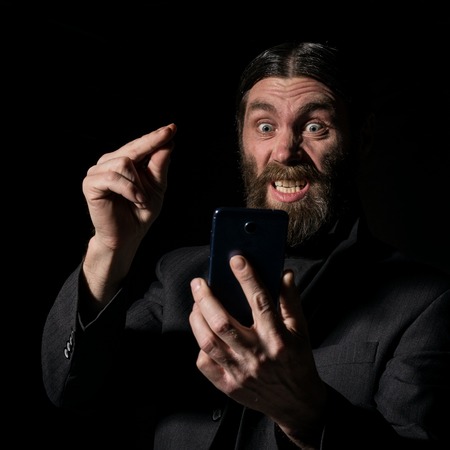 Old believer senior priest with a smartphone, bearded old man is calling on a dark backgroundの写真素材