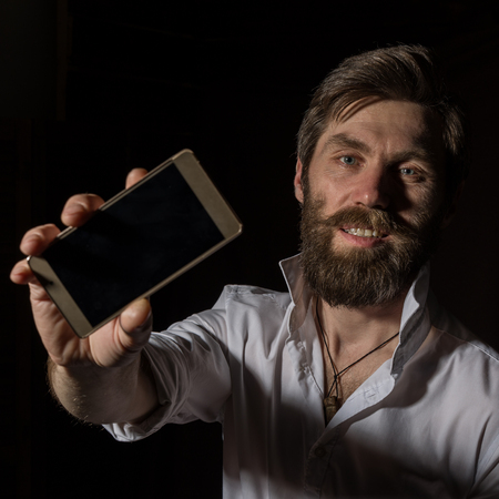 handsome bearded man shows his phone with fascinating smile on a dark background.の写真素材
