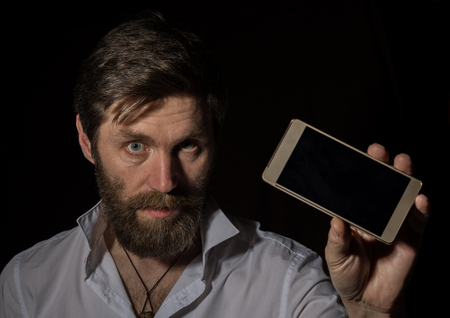 handsome bearded man shows his phone with fascinating smile on a dark background.の写真素材