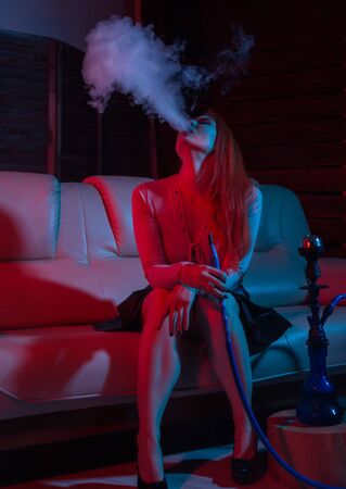 seductive redhead woman sitting on a sofa and smokes hookah in red blue tonesの写真素材