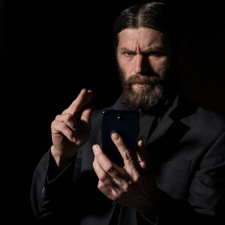 Old believer senior priest with a smartphone, bearded old man is calling on a dark backgroundの写真素材