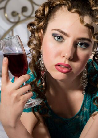beautyful young curly woman resting on the bed in a hotel room and enjoying red wineの写真素材