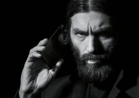 strange bearded senior priest with a smartphone, bearded old man is calling on a dark background. black and whiteの写真素材