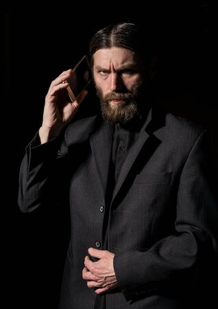 strange bearded senior priest with a smartphone, bearded old man is calling on a dark backgroundの写真素材