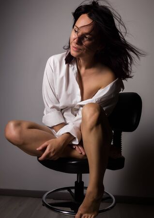Young beautiful hipster girl in a white shirt with bare shoulder sitting on chair on a gray backgroundの写真素材
