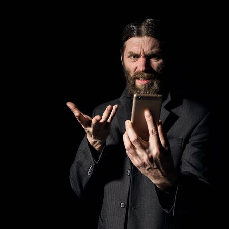 strange bearded senior priest with a smartphone, bearded old man is calling on a dark backgroundの写真素材