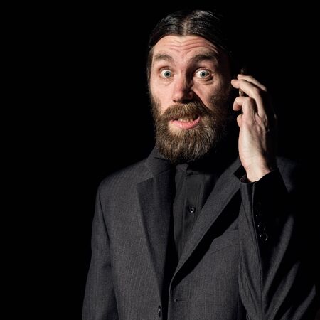 strange bearded senior priest with a smartphone, bearded old man is calling on a dark backgroundの写真素材