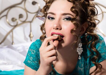 young happy cheerful curly woman lying in a bed and eating chocolateの写真素材