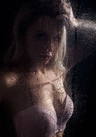 beautyful blonde woman behind wet glass on a dark background, woman takes a showerの写真素材