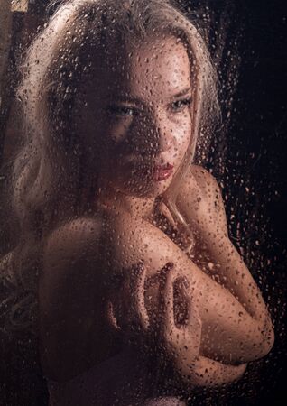 beautyful blonde woman behind wet glass on a dark background, woman takes a showerの写真素材