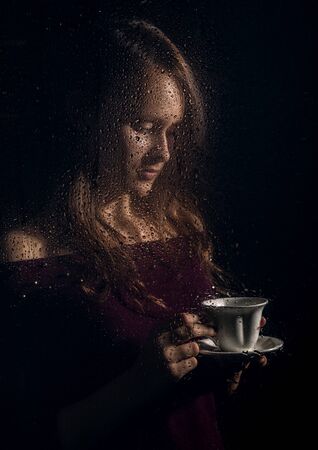 nice young girl drinks coffee behind the wet glass, raindrops on glassの写真素材