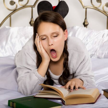 sad young student girl reads a book while lying on a bed doing homeworkの写真素材