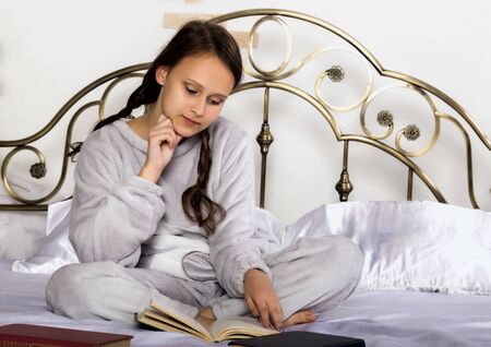 young student girl reads a book while lying on a bed doing homeworkの写真素材