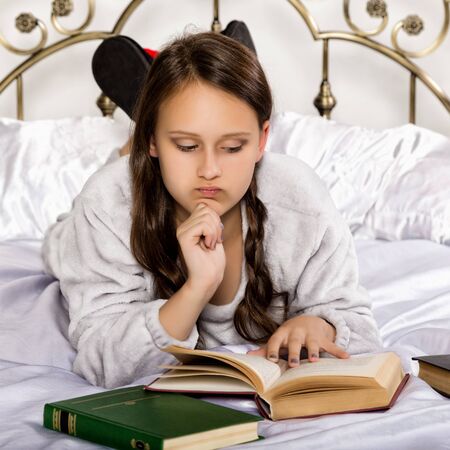 sad young student girl reads a book while lying on a bed doing homeworkの写真素材