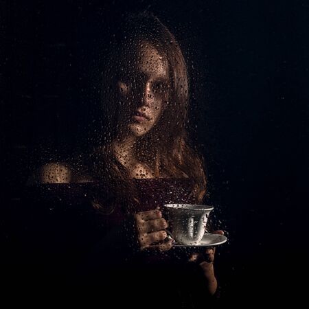 nice young girl drinks coffee behind the wet glass, raindrops on glassの写真素材