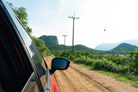 View from the car windshield from the driver's seat. Travel in spring or summer weather. Beautifull view of the car side mirror. Dirty Rural Road In Summer Field, Meadow, mountain,Countryside.の写真素材
