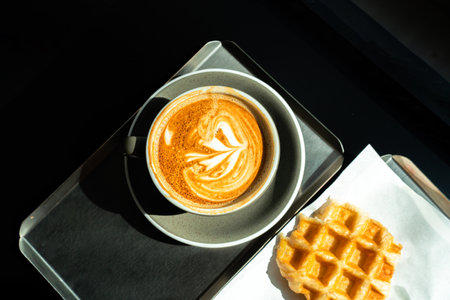 Late coffee art on the wooden table. relaxation concept. Iced coffee latte with soft white milk foam and sunlight shining through. Photograph of a cup of hot coffee and Croffle on table in a coffee/bar in during sunny day.の写真素材