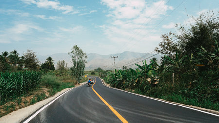 Biker is riding a motorcycle on the road ascending to the top of the hill. beautiful road and mountain scenery, Asphalt road through the green field and clouds on blue sky in summer day. Beautiful grassland road in Thailand.の写真素材