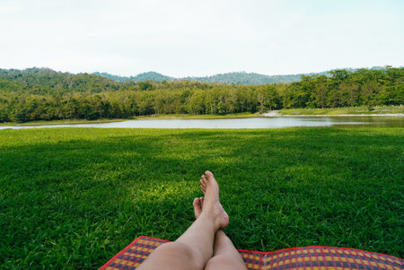 Female legs in Barefoot on picnic mat green grass look at lake and mountains. Woman relaxing on sunny day at Jedkod park, Thailandの写真素材