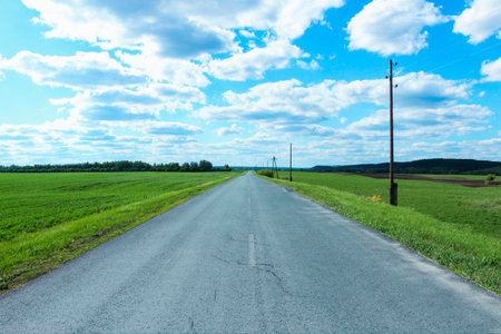 A straight asphalt country road running into the distance through fields and a sky with cloudsの写真素材
