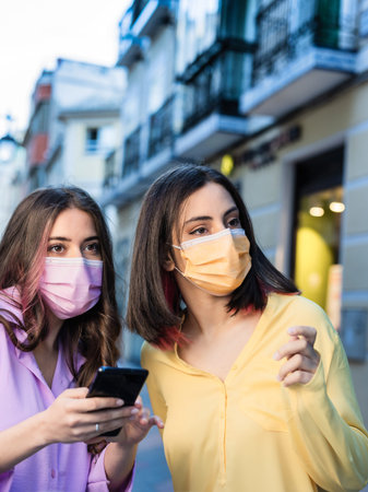 Female hispanic friends, wearing mask, search destination and shopping with smartphone. Texting and using navigator holding a mobile phone. Lifestyle portrait yellow and lavender year colorの写真素材