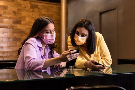 Hispanic young female friends using smartphone technology in restaurant to order dinner and...
