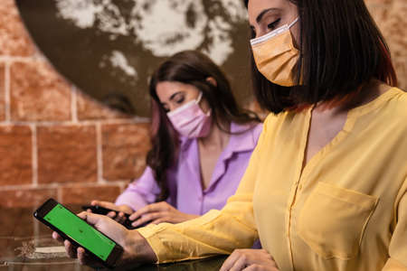 Hispanic woman using smartphone to order and pay online menu in a restaurant. New normal life with friends. Holding cellphone with chroma key green screen. Yellow color of year 2021の写真素材