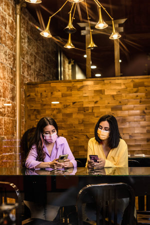 Hispanic young female friends using smartphone technology in restaurant to order dinner and chatting. Wearing face mask. Lifestyle from independent women together. Yellow and lavender color 2021の写真素材