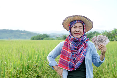 Asian farmer woman is at paddy field, hold Thai banknote money. Concept , Farmer happy to get profit, income, agriculture supporting money. Proud on crops.の写真素材