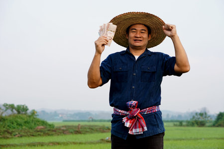 Happy Asian man farmer is at paddy field, holds Thai banknote money.  Concept : Happy farmer get profit, income, agriculture supporting money. Proud on crops.の写真素材