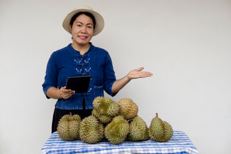 Asian woman gardener wears hat, hold smart tablet, stands beside of durian fruits.  Concept , fruit seller use internet technology device  to sell agriculture crops online. Grow duの写真素材