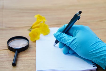 Hand in blue glove writing in notebook with magnifying glass and yellow flowerの写真素材