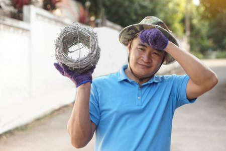Asian man worker holds roll of barbed wire, feels tired. Concept, construction tool. Barbed wire is used for make fences , secure property ,make border to show the territory areaの写真素材
