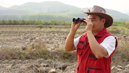 Asian man explorer wears hat, red vest shirt, holds binocular to explore boundary land. Concept,  land property exploration. Long vision distance, zoom for clearing  scene.の写真素材