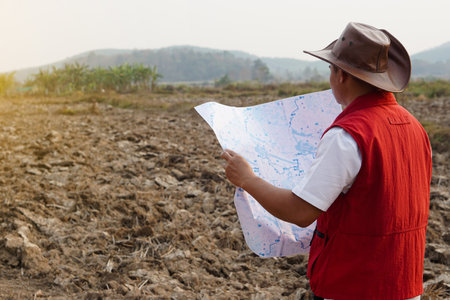 Asian man explorer wears hat, red vest shirt, holds map to explore land boundary. Concept, land planning, exploring property. Geodetic survey area. Treasure  hunting on land.の写真素材