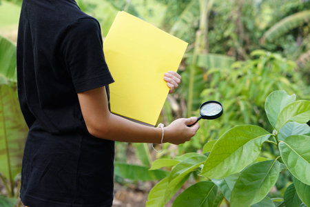 Closeup boy is learning about botanical plants lesson, holds magnifying glass and book. Concept, Science and environment project assignment from school. Self learning outside.の写真素材