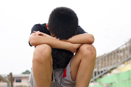 Depressed boy sits alone outdoor, sad and cry, face down between his knees. concept, emotion, feeling. Disappointed unhappy guilty . Boy is bullied and punished. Lonely.の写真素材