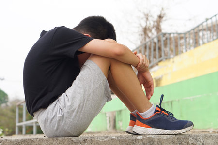 Depressed boy sits alone outdoor, sad and cry, face down between his knees. concept, emotion, feeling. Disappointed unhappy guilty . Boy is bullied and punished. Lonely.の写真素材